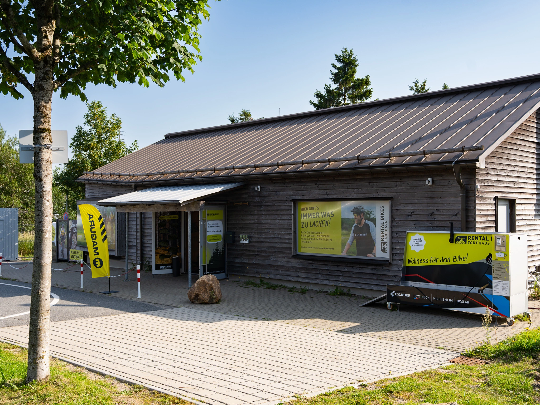 Die "Speckhütte" auf Torfhaus. Hier starten wir unsere Touren im Harz. Ein kleines Holzgebäude mit großen Fenstern, Außenschildern und einem Wasch-Automaten für den Fahrradverleih steht an einem sonnigen Tag an einem gepflasterten Gehweg, mit Bäumen und Grün im Hintergrund.