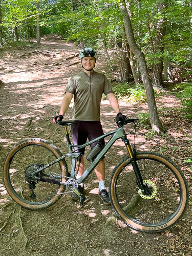 image1-1-3 Eine Person, die einen Helm und eine Fahrradausrüstung trägt, steht lächelnd mit einem Mountainbike auf einem Waldweg, umgeben von Bäumen und Sonnenlicht, das durch die Blätter fällt.